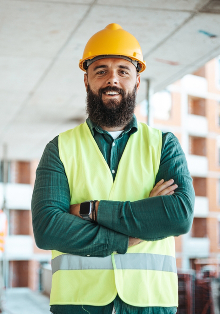 A smiling male construction worker in a yellow hard hat and reflective vest stands with arms crossed at a busy site, with colleagues in the background.